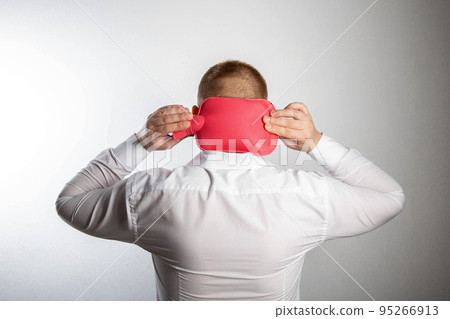 A man holds a heating pad with hot water on the back of his head. Muscle relaxation and vasodilatation of the head, thermotherapy. Headache treatment, close-up A man holds a heating pad with hot water on the back of his head. Muscle relaxation and vasodilatation of the head, thermotherapy. Headache treatment, close-up 95266913