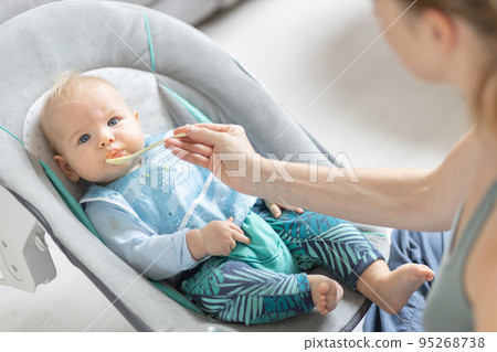 Mother spoon feeding her baby boy infant child in baby chair with fruit puree. Baby solid food introduction concept. Mother spoon feeding her baby boy infant child in baby chair with fruit puree. Baby solid food introduction concept. 95268738