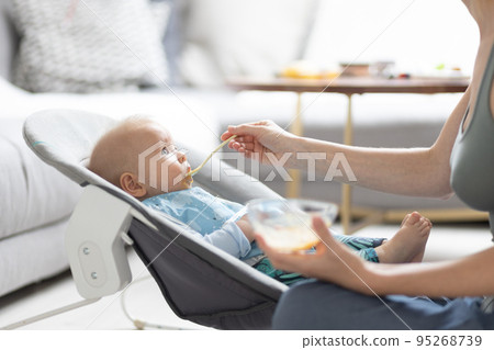 Mother spoon feeding her baby boy infant child in baby chair with fruit puree. Baby solid food introduction concept. 95268739