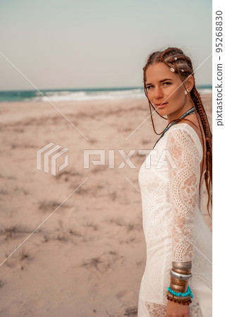 Model in boho style in a white long dress and silver jewelry on the beach. Her hair is braided, and there are many bracelets on her arms. 95268830