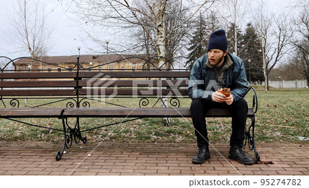 Hungry bearded homeless man sits on a bench and eats a bun in a city park. Below poverty line. Male tramp in dirty clothes with cap and hat. Beggar. Illegal immigrant. Drunkenness, alcoholism 95274782