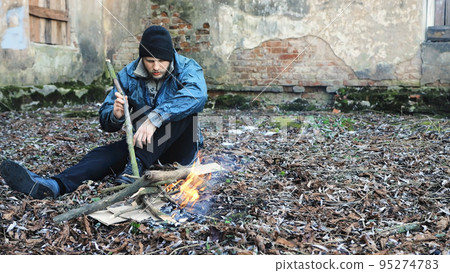 Homeless man sits on ground and uses stick to keep fire burning to keep warm from cold. Tramp in dirty clothes and hat cap. Below poverty line. Illegal immigrant. Male refugee. Fugitive beggar Homeless man sits on ground and uses stick to keep fire burning to keep warm from cold. Tramp in dirty clothes and hat cap. Below poverty line. Illegal immigrant. Male refugee. Fugitive beggar 95274783