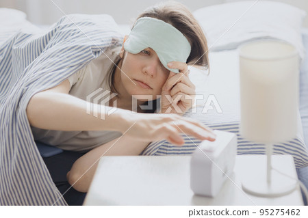 woman in bed with a sleep mask wakes up and turns off the alarm. a clock with a timer, a woman yawns early in the morning woman in bed with a sleep mask wakes up and turns off the alarm. a clock with a timer, a woman yawns early in the morning 95275462