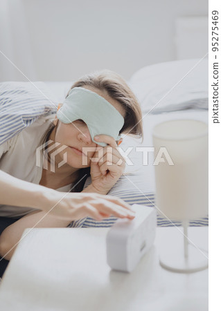 woman in bed with a sleep mask wakes up and turns off the alarm. a clock with a timer, a woman yawns early in the morning woman in bed with a sleep mask wakes up and turns off the alarm. a clock with a timer, a woman yawns early in the morning 95275469