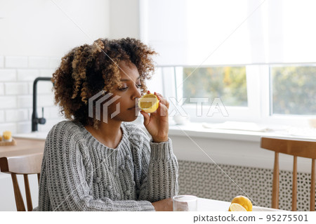 African American woman with a cold sniffs lemon to check her well-being, measures her body temperature African American woman with a cold sniffs lemon to check her well-being, measures her body temperature 95275501