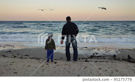 Two people an elderly man and small child stand on the seashore and feed seagulls in the evening. 95275779