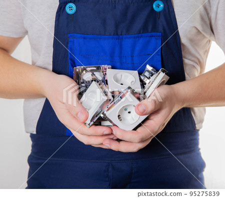 A man in blue overalls holds a lot of electrical sockets in his palms. The concept of electrician services, installation and connection of sockets, close-up A man in blue overalls holds a lot of electrical sockets in his palms. The concept of electrician services, installation and connection of sockets, close-up 95275839