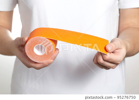 The girl holds a roll of orange kinesiology tape in her hands on the background of a white T-shirt. Close-up The girl holds a roll of orange kinesiology tape in her hands on the background of a white T-shirt. Close-up 95276750