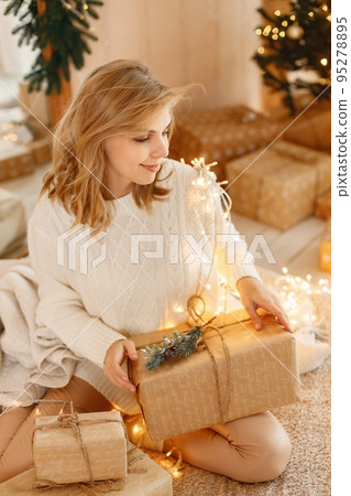 Portrait of a woman celebrating Christmas alone. Girl sitting wrapped in Christmas lights holding gift boxes and posing for a photo. Girl wearing beige knitted clothes. 95278895