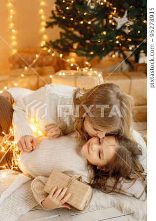 Young mother and her daughter lay on a floor in Christmas atmosphere. Girl and her mother having fun wrapping in Christmas lights. Family wearing beige knitted clothes. 95278915