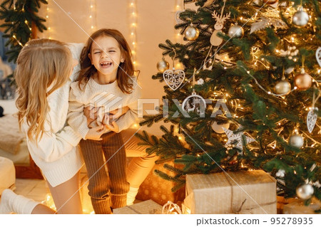 Portrait of a little girl celebrating Christmas with her mom at home. Girl and her mother having fun while standing near Christmas tree. Family wearing beige knitted clothes. 95278935