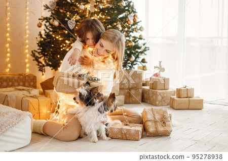 Portrait of a little girl celebrating Christmas with her dogs. Girl and her mother sitting near Christmas tree and posing for a photo. Family wearing beige knitted clothes. 95278938