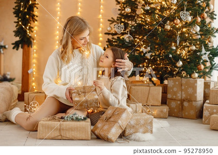 Portrait of a little girl celebrating Christmas and holding a gift box. Girl and her mother sitting near Christmas tree and posing for a photo. Family wearing beige knitted clothes. Portrait of a little girl celebrating Christmas and holding a gift box. Girl and her mother sitting near Christmas tree and posing for a photo. Family wearing beige knitted clothes. 95278950