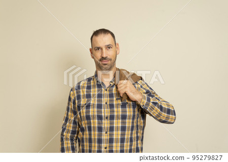 Latin handsome bearded man posing in a checkered shirt with a brown jacket on his shoulder, isolated over beige background. 95279827