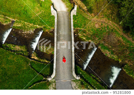 Stone arched bridge in the park, pedestrian bridge over the cascading river, woman lying on the bridge, aerial view. Stone arched bridge in the park, pedestrian bridge over the cascading river, woman lying on the bridge, aerial view. 95281800