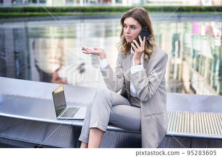 Disappointed businesswoman talking on mobile phone, sitting with laptop and shrugging clueless. 95283605