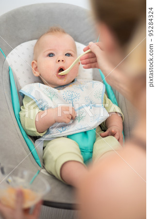 Mother spoon feeding her baby boy infant child in baby chair with fruit puree. Baby solid food introduction concept. 95284324