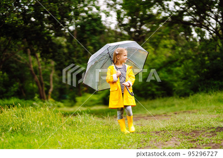 Little girl with transparent umbrella playing in the rain on sunny. Beautiful girl in a yellow cloak and rubber boots. 95296727