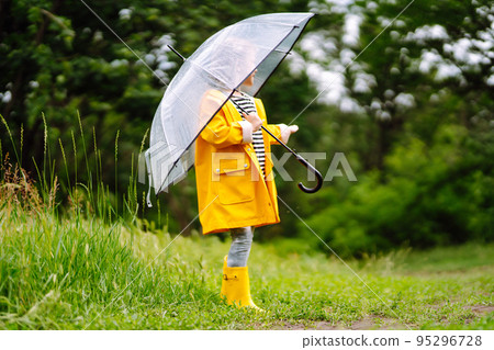 Little girl with transparent umbrella playing in the rain on sunny. Beautiful girl in a yellow cloak and rubber boots. 95296728