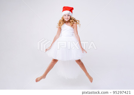 Little joyful girl in red Santa hat and white beautiful dress jumps from Little joyful girl in red Santa hat and white beautiful dress jumps from 95297421