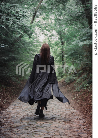 A beautiful woman in a black raincoat walks alone through a dense forest, back view 95299668