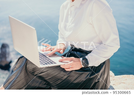 Digital nomad, Business woman working on laptop by the sea. Pretty lady typing on computer by the sea at sunset, makes a business transaction online from a distance. Freelance, remote work on vacation 95300665