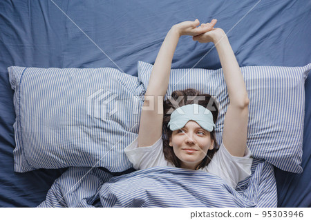 top view of a woman in bed wearing a sleep mask, a happy European woman wakes up and stretches, does a warm-up top view of a woman in bed wearing a sleep mask, a happy European woman wakes up and stretches, does a warm-up 95303946
