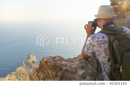 A young man with a backpack takes pictures of the landscape on top of a mountain. Back view A young man with a backpack takes pictures of the landscape on top of a mountain. Back view 95305345