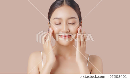 Face cream commercial advertising concept. Young beautiful Asian woman massages her face gently with fingertips isolated over beige brown background. 95308401