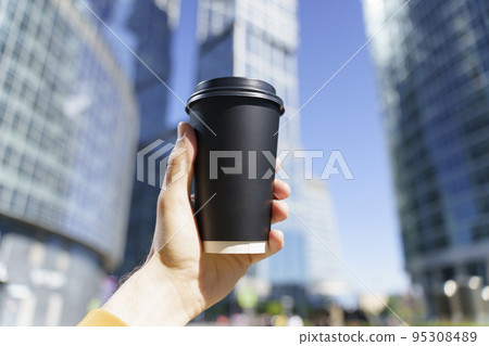 Takeaway coffee in a paper cup. A hand with a drink to go on the background of a busy city street. Paper cup mockup 95308489