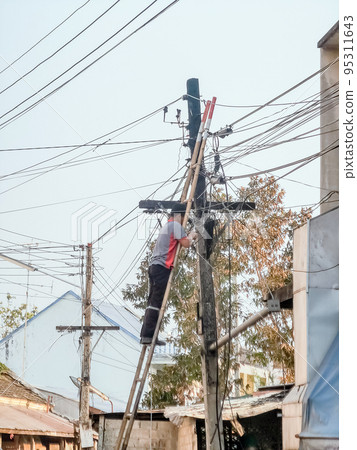 Asian electrician on a ladder repairing wire of the power line on electric power pole in the village. 95311643