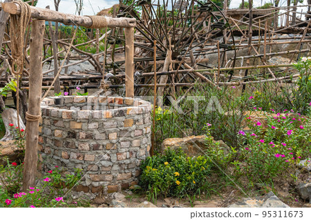Beautiful artesian well made by bricks with cement and wheel pulley with metal bucket and rope in peaceful garden atmosphere. Retro stone water well in rural area.Garden decoration with antique items 95311673