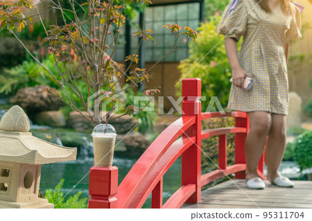 Iced cappuccino with straws placed on head of red wooden bridge with blurred image of woman standing on bridge and small waterfalls and cool flowing pond in garden decorated in Japanese style. Iced cappuccino with straws placed on head of red wooden bridge with blurred image of woman standing on bridge and small waterfalls and cool flowing pond in garden decorated in Japanese style. 95311704