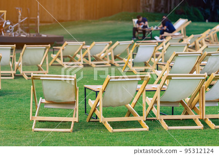Many empty white deck chairs with tables in lawn is surrounded by shady green grass. Comfortable on outdoor patio chairs in garden. Lawn chairs in the park. Sunbeds in the garden. Selective focus. Many empty white deck chairs with tables in lawn is surrounded by shady green grass. Comfortable on outdoor patio chairs in garden. Lawn chairs in the park. Sunbeds in the garden. Selective focus. 95312124