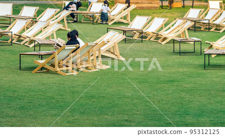 Asian young couple sit to relax and look at the menu to order food for dinner on white deck chairs with tables in lawn is surrounded by shady green grass.Comfortable on outdoor patio chairs in garden. 95312125
