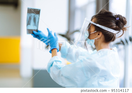 A female doctor in visor and protective gloves examines an X-ray of a patient 95314117