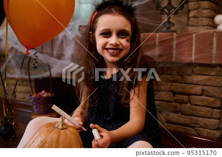 Mischievous child, a little girl dressed as a witch with a spider painted on her face, smiles looking at camera, drawing a scary face on a pumpkin, a Jack-O-Lantern. Concept of a happy Halloween party Mischievous child, a little girl dressed as a witch with a spider painted on her face, smiles looking at camera, drawing a scary face on a pumpkin, a Jack-O-Lantern. Concept of a happy Halloween party 95315170
