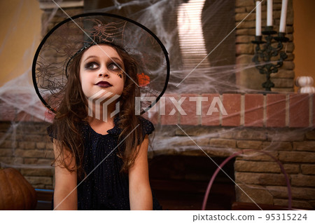 Portrait of little girl wearing a witch carnival costume and wizard hat, with face art makeup, a black spider paint on her cheek, sitting against a cobweb-covered fireplace. Halloween festival concept Portrait of little girl wearing a witch carnival costume and wizard hat, with face art makeup, a black spider paint on her cheek, sitting against a cobweb-covered fireplace. Halloween festival concept 95315224