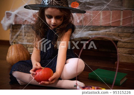 Adorable little girl looking like a sorceress playing with a bright orange pumpkin, sitting on the floor near a spell book, against a spider web covered fireplace. Halloween party concept Adorable little girl looking like a sorceress playing with a bright orange pumpkin, sitting on the floor near a spell book, against a spider web covered fireplace. Halloween party concept 95315226
