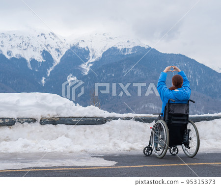 A woman in a wheelchair holds her hands in the shape of a heart in the mountains in winter. A woman in a wheelchair holds her hands in the shape of a heart in the mountains in winter. 95315373