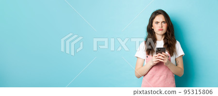 Disappointed girl frowning and grimacing displeased after using smartphone app, holding phone and express dislike, standing over blue background 95315806