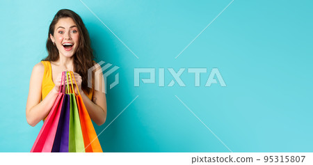 Portrait of surprised young woman look amazed at camera, holding shopping bags, see discounts in store, standing over blue background 95315807
