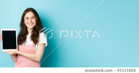 Portrait of smiling beautiful woman demonstrate blank tablet screen, showing promo on device screen, standing against blue background 95315808