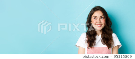 Close up of dreamy silly girl thinking of something, looking at upper left corner and smiling happy, daydreaming, staring at logo, standing over blue background 95315809