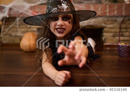 Scary child girl, a sorceress in a wizard's hat, stretches out her hand, looks at the camera with a frightening look, lies on the floor near a brick wall covered with cobwebs, and Halloween decoration Scary child girl, a sorceress in a wizard's hat, stretches out her hand, looks at the camera with a frightening look, lies on the floor near a brick wall covered with cobwebs, and Halloween decoration 95316190