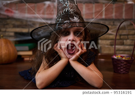 Funny child girl in wizard's hat, a little enchantress, lies on the floor, casts spells looking at camera, surrounded by a fireplace covered with spider web, pumpkins, Halloween treat and sorcery book Funny child girl in wizard's hat, a little enchantress, lies on the floor, casts spells looking at camera, surrounded by a fireplace covered with spider web, pumpkins, Halloween treat and sorcery book 95316191
