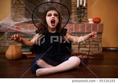 A gothic mysterious child, a little girl witch or sorceress wearing a black dress and wizard's hat, casts spells while sitting on the floor by a fireplace with cobwebs pumpkins and Halloween treats. 95316193