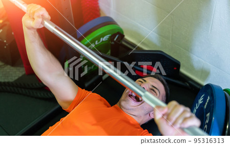 Amazing image of a young man doing bench press with a maximum weight. He screams in the effort. Exciting and motivational photo. Amazing image of a young man doing bench press with a maximum weight. He screams in the effort. Exciting and motivational photo. 95316313