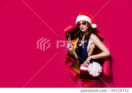 Young, sexy, beautiful woman in swimsuit, Santa hat, holding gift box, glass of champagne peeking out of a hole in a red studio paper background. Christmas, xmas, x-mas and winter concept Young, sexy, beautiful woman in swimsuit, Santa hat, holding gift box, glass of champagne peeking out of a hole in a red studio paper background. Christmas, xmas, x-mas and winter concept 95316712