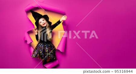 a little girl in a witch costume peeks out of a hole in a paper background 95316944